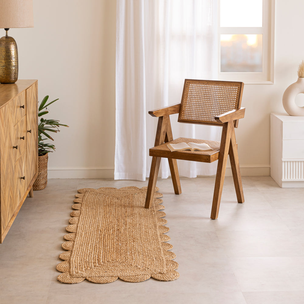 A hand-braided natural jute runner rug with a scalloped edge laid on a light beige floor in a modern room, placed alongside a chevron-patterned wooden sideboard and a cane armchair.
