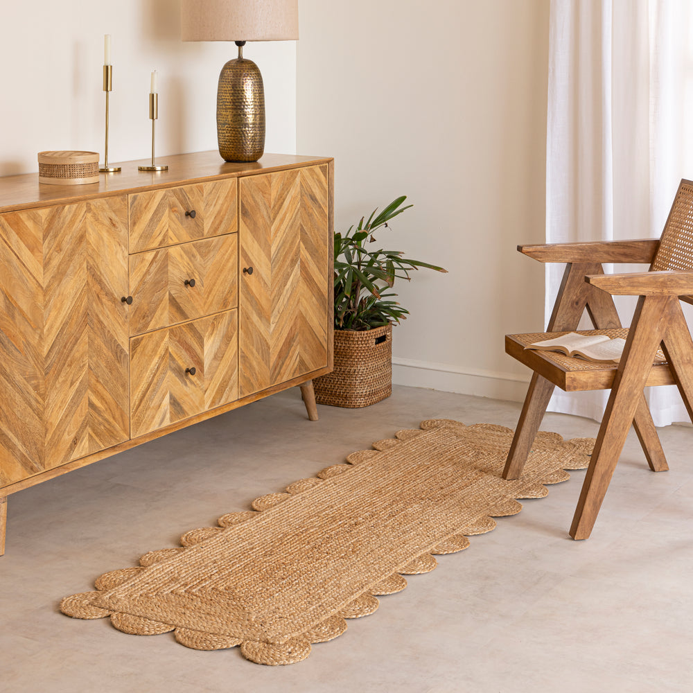 A hand-braided natural jute runner rug with a scalloped edge laid on a light beige floor in a modern room, placed alongside a chevron-patterned wooden sideboard and a cane armchair.
