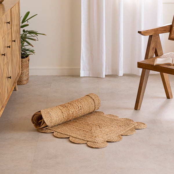 A hand-braided natural jute runner rug with a scalloped edge laid on a light beige floor in a modern room, placed alongside a chevron-patterned wooden sideboard and a cane armchair.