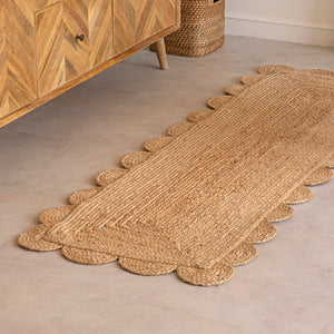 A hand-braided natural jute runner rug with a scalloped edge laid on a light beige floor in a modern room, placed alongside a chevron-patterned wooden sideboard and a cane armchair.