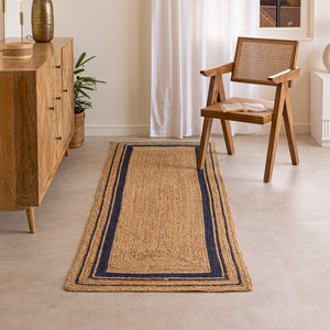 Handwoven natural jute runner rug with navy blue geometric border, styled in a warm modern living room with wooden furniture and a cane chair.
