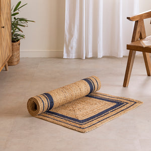 Handwoven natural jute runner rug with navy blue geometric border, styled in a warm modern living room with wooden furniture and a cane chair.