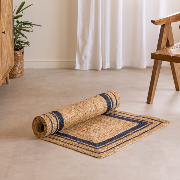 Handwoven natural jute runner rug with navy blue geometric border, styled in a warm modern living room with wooden furniture and a cane chair.