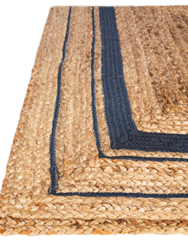 Natural woven jute rug with a navy blue double-border design, styled on a rustic wooden floor beside a wooden bench, striped throw blanket, and green potted fern.