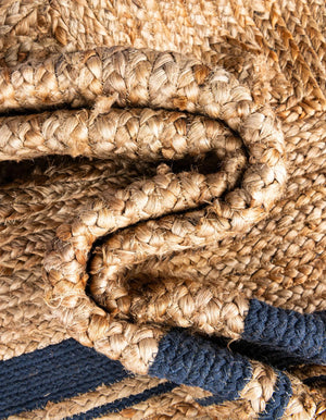 Natural woven jute rug with a navy blue double-border design, styled on a rustic wooden floor beside a wooden bench, striped throw blanket, and green potted fern.