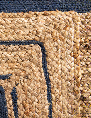 Natural woven jute rug with a navy blue double-border design, styled on a rustic wooden floor beside a wooden bench, striped throw blanket, and green potted fern.
