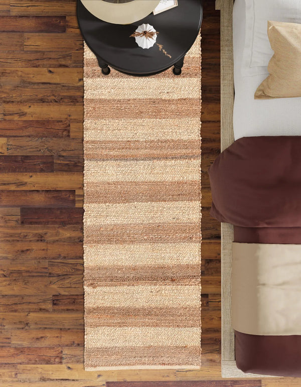 A bright, rustic entryway featuring a long natural jute runner rug laid over light wooden flooring. To the right sits a distressed white wooden bench with a linen throw blanket draped over it, a decorative ceramic vase on top, and woven storage baskets tucked underneath. Nearby, a potted fiddle-leaf fig plant rests in a terracotta pot next to a white door, with a round woven mirror hanging on the wall above the bench.