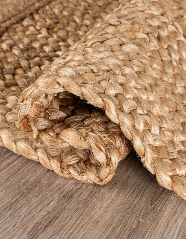 Large rectangular handwoven natural jute area rug with a thick chunky braided weave, styled under a white sofa and a rustic wooden coffee table in a warm minimalist living room.