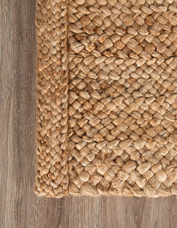 Large rectangular handwoven natural jute area rug with a thick chunky braided weave, styled under a white sofa and a rustic wooden coffee table in a warm minimalist living room.