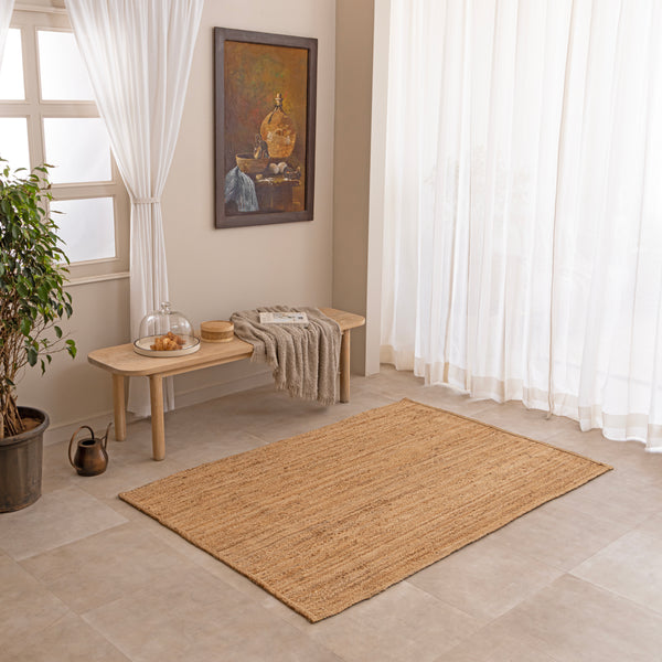Handwoven natural jute area rug in warm beige and earthy tones, featuring a textured woven design styled in a cozy neutral room with a vase and book.
