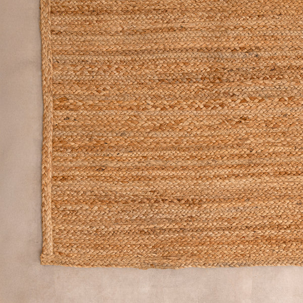 Handwoven natural jute area rug in warm beige and earthy tones, featuring a textured woven design styled in a cozy neutral room with a vase and book.