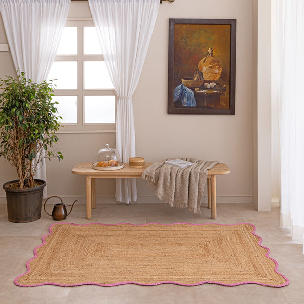 Handcrafted natural jute area rug featuring a diamond center weave and a vibrant pink scalloped border, placed on a tiled floor in a modern boho room setting.