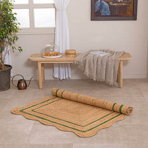 Handcrafted natural jute area rug with a double green border line and a wavy scalloped edge, displayed on a beige tile floor.