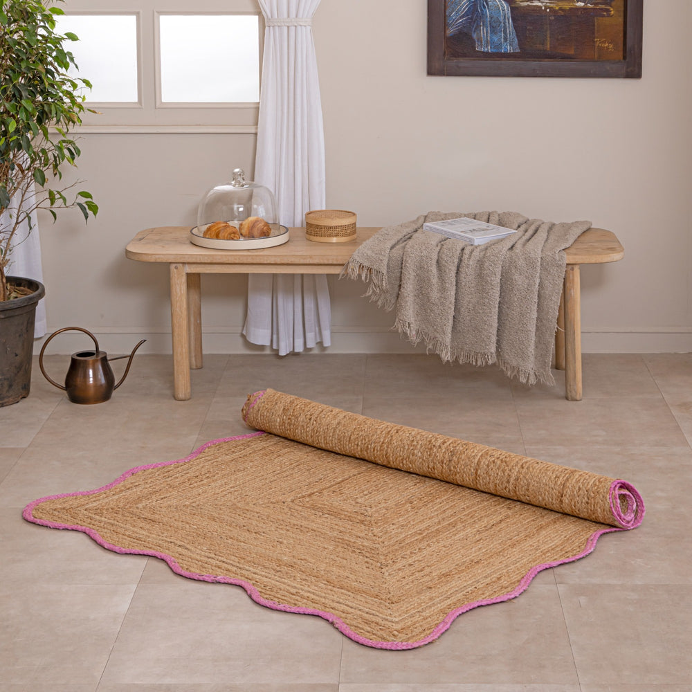 Handcrafted natural jute area rug featuring a diamond center weave and a vibrant pink scalloped border, placed on a tiled floor in a modern boho room setting.