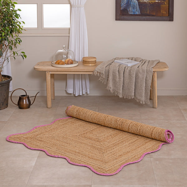 Handcrafted natural jute area rug featuring a diamond center weave and a vibrant pink scalloped border, placed on a tiled floor in a modern boho room setting.