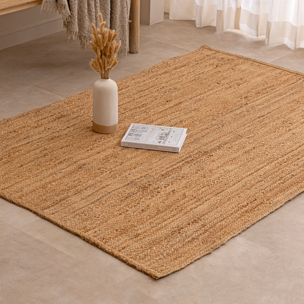 Handwoven natural jute area rug in warm beige and earthy tones, featuring a textured woven design styled in a cozy neutral room with a vase and book.