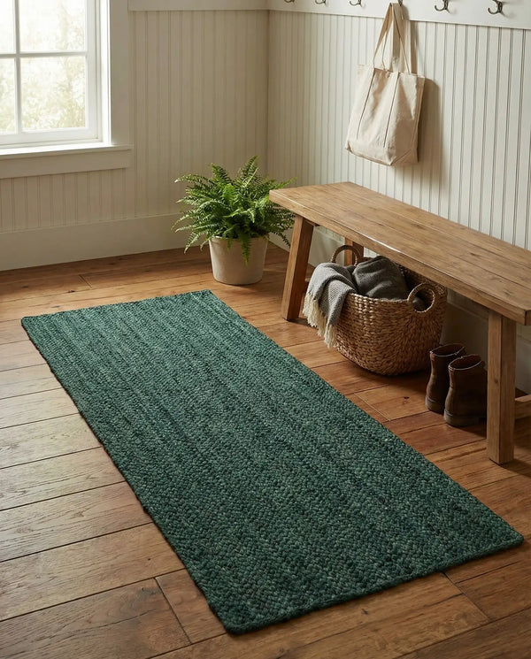 Deep green handwoven braided jute runner rug placed on a hardwood floor alongside a wooden bench in an entryway.