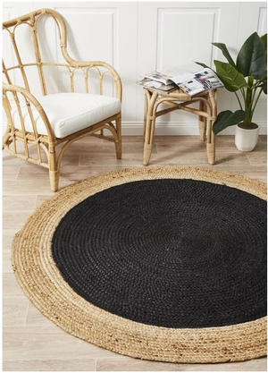 A high-angle view of a modern living room featuring a round braided jute area rug with a large charcoal black center and a natural tan border, placed on light hardwood flooring in front of a beige sofa and wooden armchair.