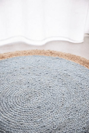 A high-angle view of a modern living room featuring a round braided jute area rug with a pastel light blue center and a natural tan border, styled on light hardwood floors with a cream sofa and a wooden armchair.