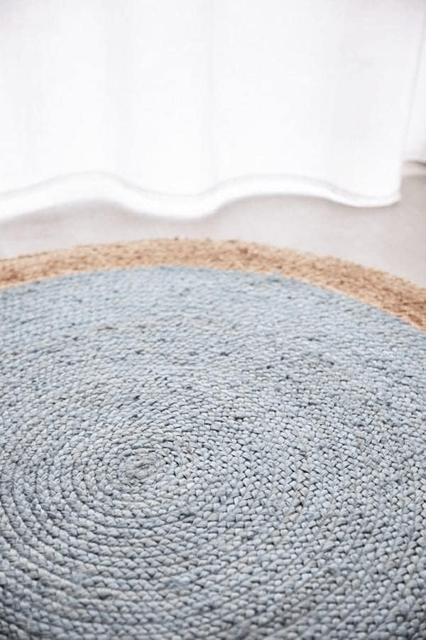 A high-angle view of a modern living room featuring a round braided jute area rug with a pastel light blue center and a natural tan border, styled on light hardwood floors with a cream sofa and a wooden armchair.
