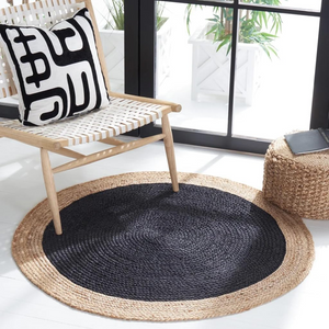 A high-angle view of a modern living room featuring a round braided jute area rug with a large charcoal black center and a natural tan border, placed on light hardwood flooring in front of a beige sofa and wooden armchair.