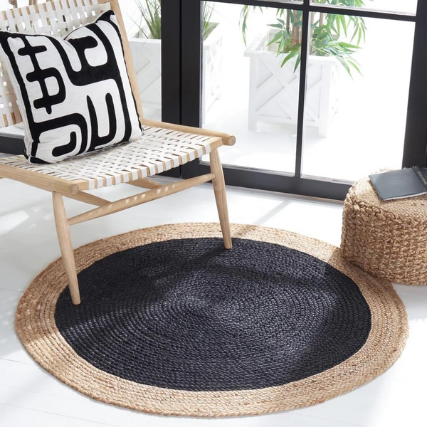 A high-angle view of a modern living room featuring a round braided jute area rug with a large charcoal black center and a natural tan border, placed on light hardwood flooring in front of a beige sofa and wooden armchair.