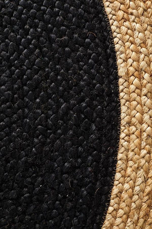 A high-angle view of a modern living room featuring a round braided jute area rug with a large charcoal black center and a natural tan border, placed on light hardwood flooring in front of a beige sofa and wooden armchair.