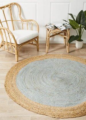 A high-angle view of a modern living room featuring a round braided jute area rug with a pastel light blue center and a natural tan border, styled on light hardwood floors with a cream sofa and a wooden armchair.