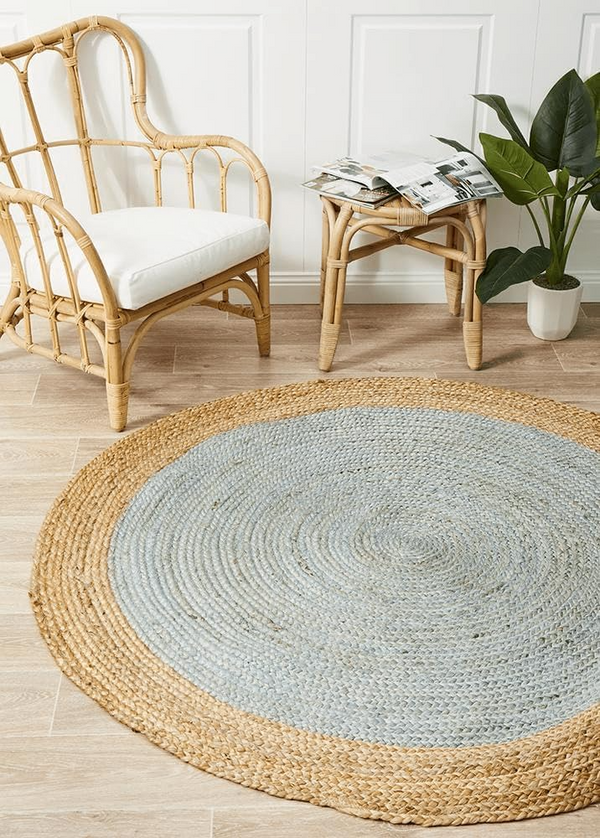 A high-angle view of a modern living room featuring a round braided jute area rug with a pastel light blue center and a natural tan border, styled on light hardwood floors with a cream sofa and a wooden armchair.