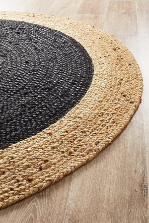 A high-angle view of a modern living room featuring a round braided jute area rug with a large charcoal black center and a natural tan border, placed on light hardwood flooring in front of a beige sofa and wooden armchair.