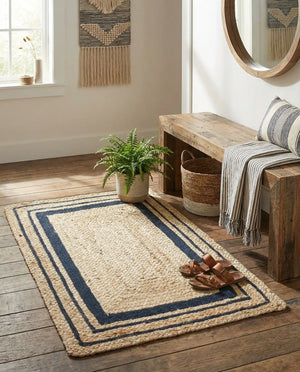 Natural woven jute rug with a navy blue double-border design, styled on a rustic wooden floor beside a wooden bench, striped throw blanket, and green potted fern.