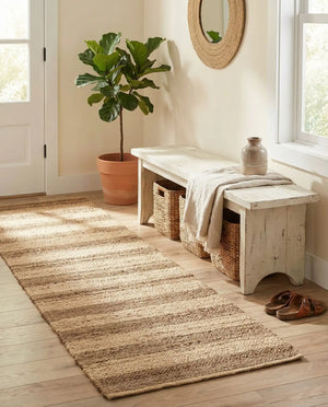 A bright, rustic entryway featuring a long natural jute runner rug laid over light wooden flooring. To the right sits a distressed white wooden bench with a linen throw blanket draped over it, a decorative ceramic vase on top, and woven storage baskets tucked underneath. Nearby, a potted fiddle-leaf fig plant rests in a terracotta pot next to a white door, with a round woven mirror hanging on the wall above the bench.