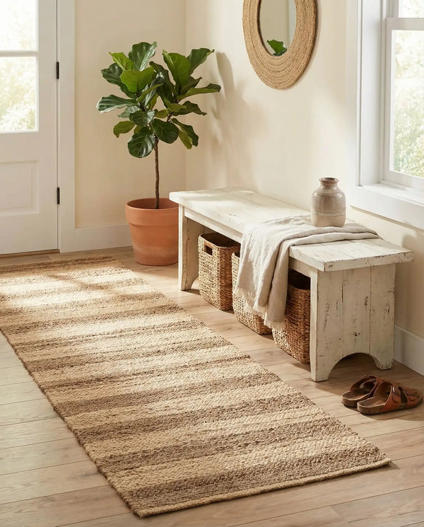 A bright, rustic entryway featuring a long natural jute runner rug laid over light wooden flooring. To the right sits a distressed white wooden bench with a linen throw blanket draped over it, a decorative ceramic vase on top, and woven storage baskets tucked underneath. Nearby, a potted fiddle-leaf fig plant rests in a terracotta pot next to a white door, with a round woven mirror hanging on the wall above the bench.
