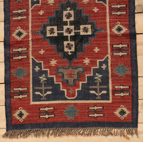 Handwoven red and navy blue tribal wool area rug with geometric Southwestern pattern and fringe, displayed on a natural wooden floor.