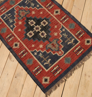 Handwoven red and navy blue tribal wool area rug with geometric Southwestern pattern and fringe, displayed on a natural wooden floor.