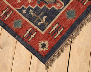 Handwoven red and navy blue tribal wool area rug with geometric Southwestern pattern and fringe, displayed on a natural wooden floor.