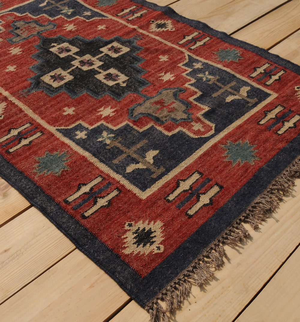 Handwoven red and navy blue tribal wool area rug with geometric Southwestern pattern and fringe, displayed on a natural wooden floor.