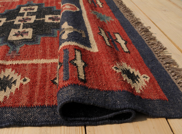 Handwoven red and navy blue tribal wool area rug with geometric Southwestern pattern and fringe, displayed on a natural wooden floor.