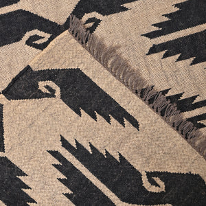 Handwoven black and ivory geometric kilim wool jute area rug in a modern living room with wooden chair and side table.
