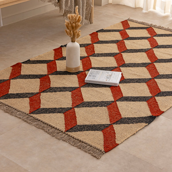 Handwoven jute and wool area rug featuring a 3D geometric optical cube pattern in terracotta rust, charcoal black, and natural beige, displayed on a tile floor with a ceramic vase and book.
