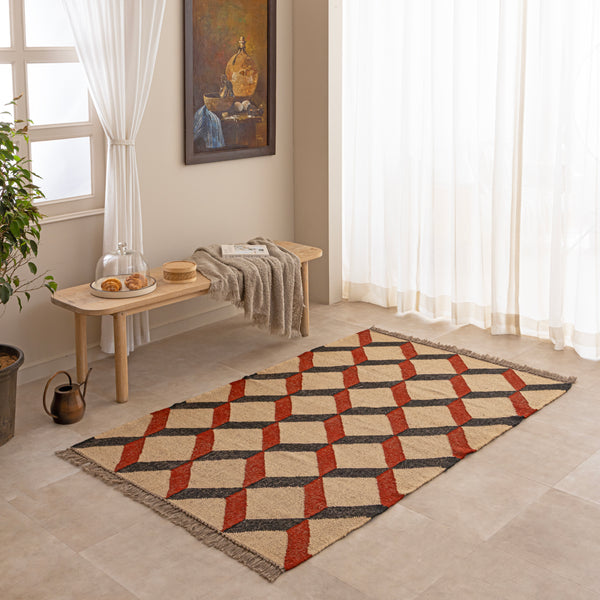 Handwoven jute and wool area rug featuring a 3D geometric optical cube pattern in terracotta rust, charcoal black, and natural beige, displayed on a tile floor with a ceramic vase and book.