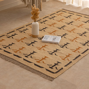 Handwoven mustard yellow and off-white geometric kilim jute rug with fringe trim, laid flat on a tile floor with a white ceramic vase and book lifestyle setting.