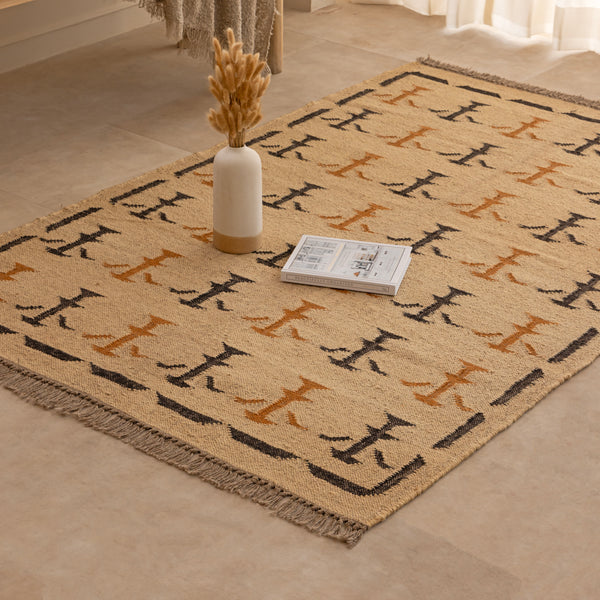 Handwoven mustard yellow and off-white geometric kilim jute rug with fringe trim, laid flat on a tile floor with a white ceramic vase and book lifestyle setting.