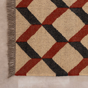 Handwoven jute and wool area rug featuring a 3D geometric optical cube pattern in terracotta rust, charcoal black, and natural beige, displayed on a tile floor with a ceramic vase and book.