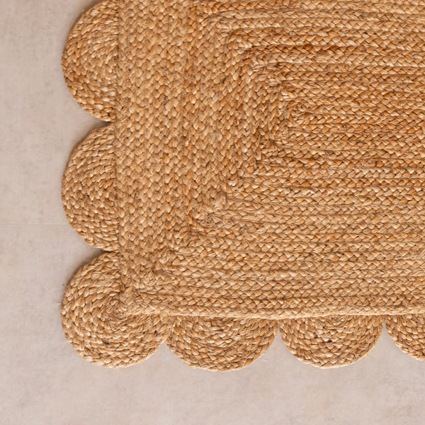 A hand-braided natural jute runner rug with a scalloped edge laid on a light beige floor in a modern room, placed alongside a chevron-patterned wooden sideboard and a cane armchair.