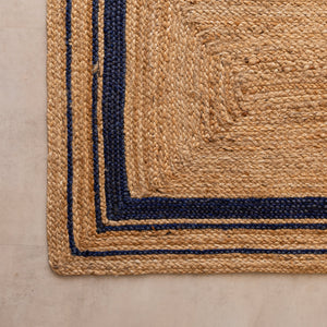 Handwoven natural jute runner rug with navy blue geometric border, styled in a warm modern living room with wooden furniture and a cane chair.