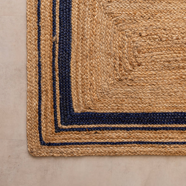 Handwoven natural jute runner rug with navy blue geometric border, styled in a warm modern living room with wooden furniture and a cane chair.