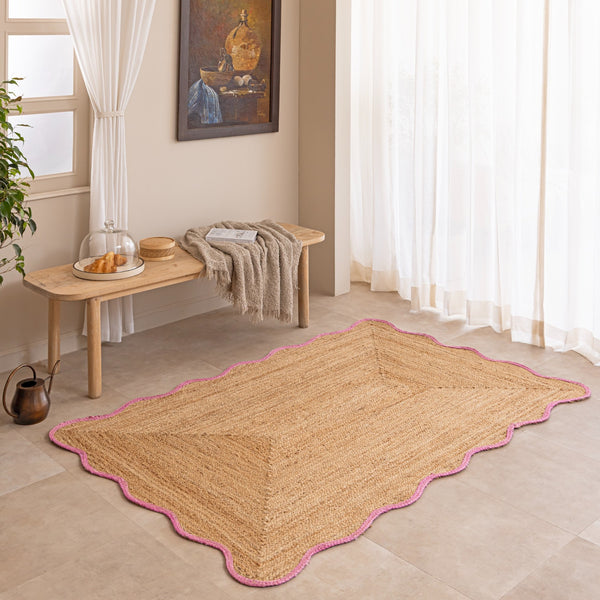 Handcrafted natural jute area rug featuring a diamond center weave and a vibrant pink scalloped border, placed on a tiled floor in a modern boho room setting.