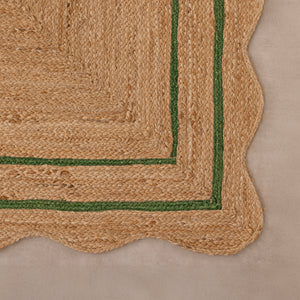 Handcrafted natural jute area rug with a double green border line and a wavy scalloped edge, displayed on a beige tile floor.