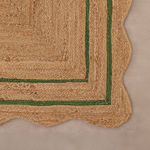 Handcrafted natural jute area rug with a double green border line and a wavy scalloped edge, displayed on a beige tile floor.
